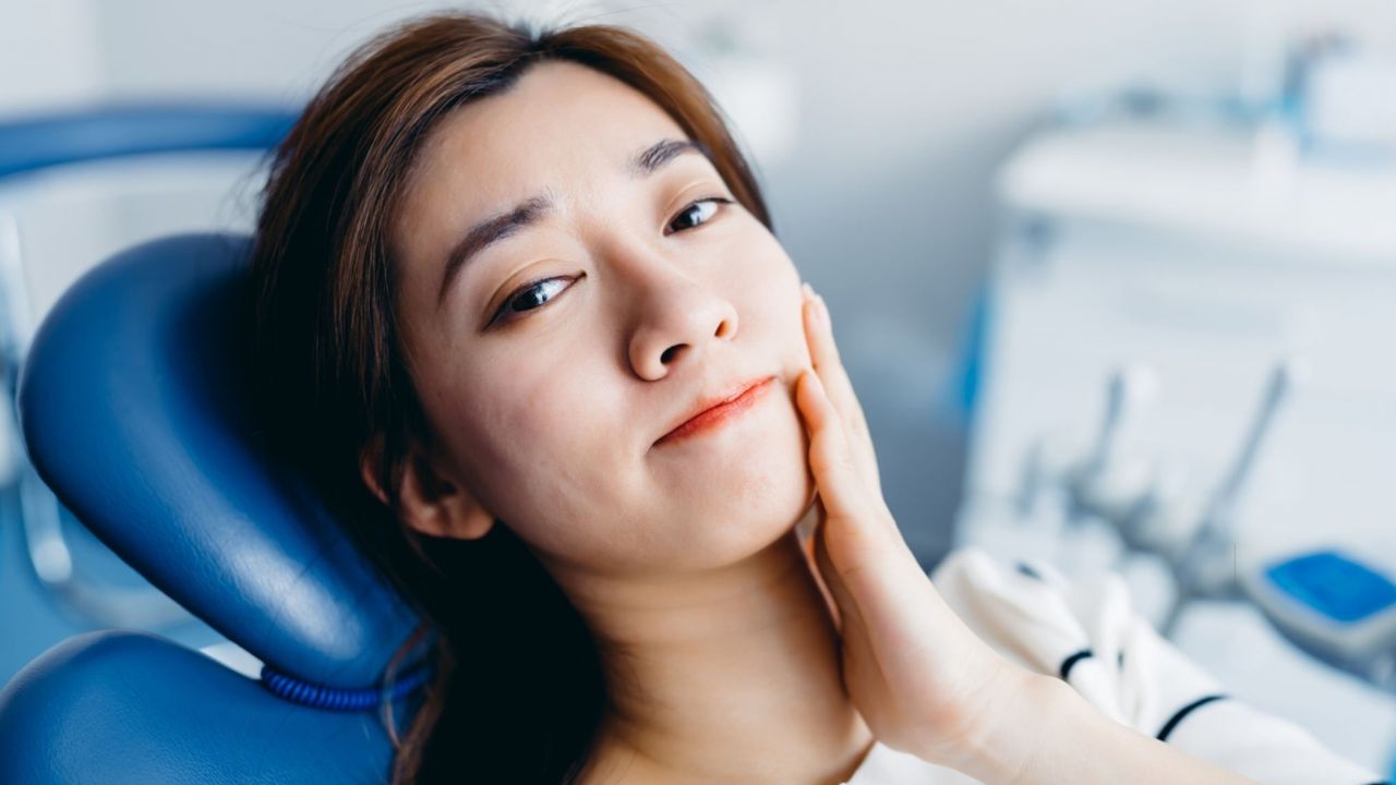 Mulher deitada em maca de dentista com mão na bochecha com queixa de dor por dtm