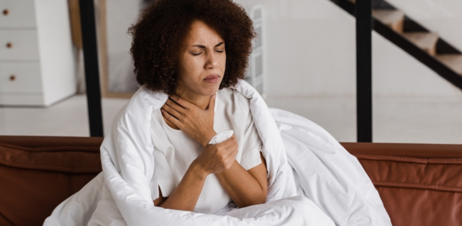 Uma mulher negra sentada com uma colcha ao redor segurando sua garganta com as duas mãos. Sua face expressa dor.
