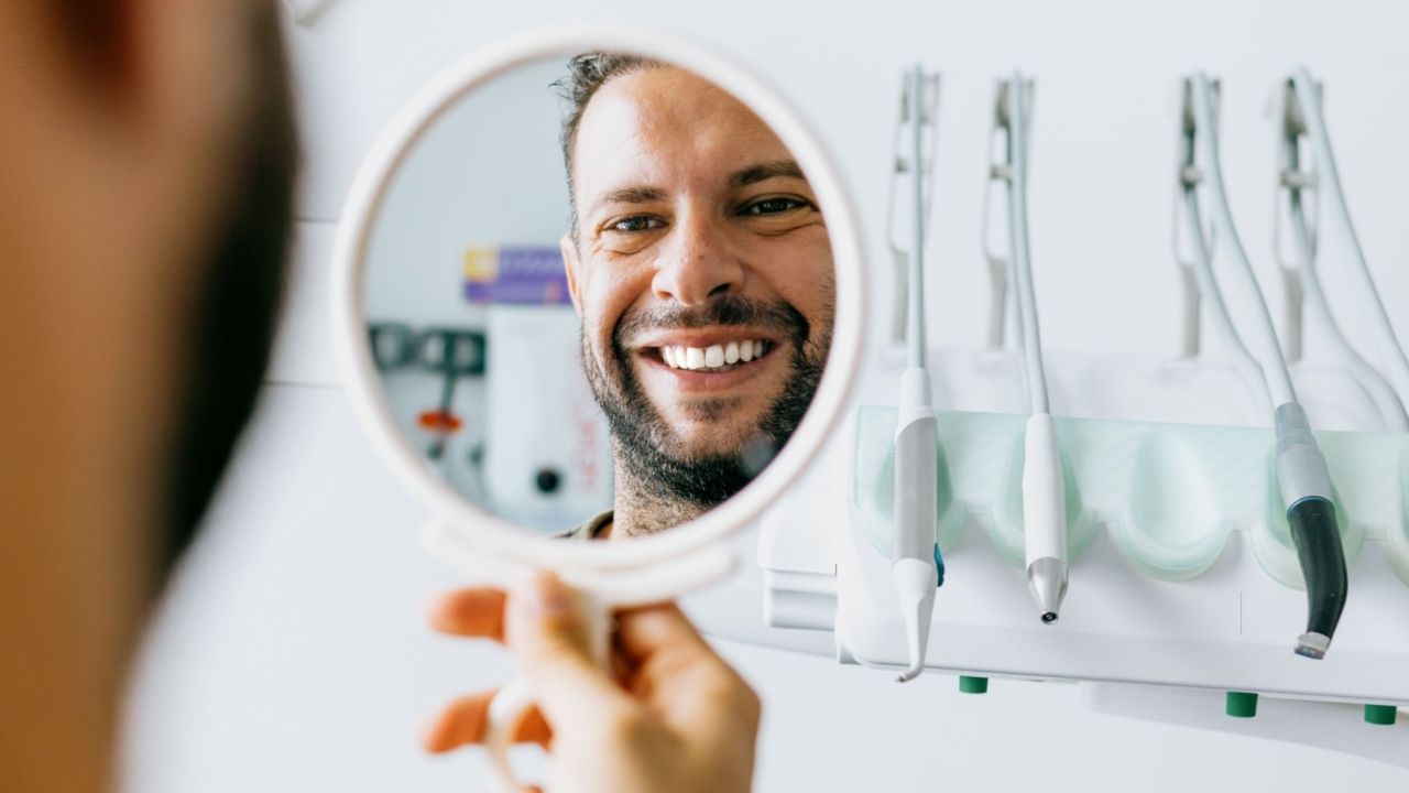 Homem sorrindo para espelho em consultório de dentista após procedimento