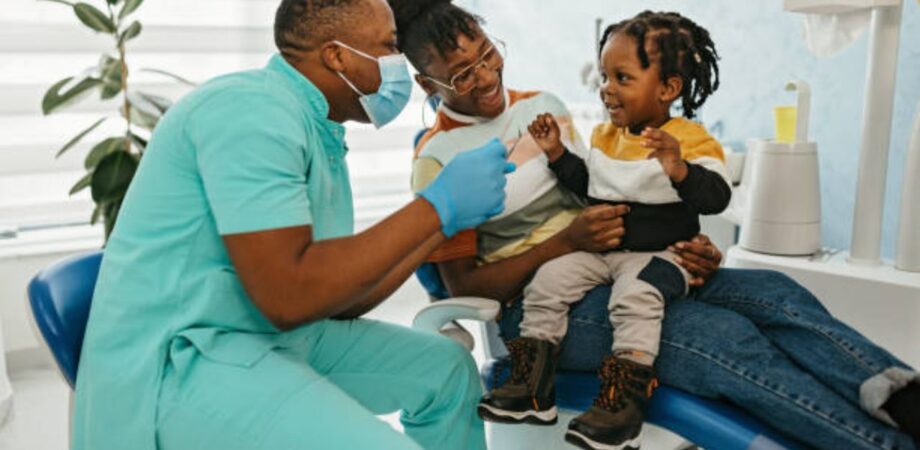 Odontopediatra atendendo criança menino que está sentado em colo da mãe em maca do consultório